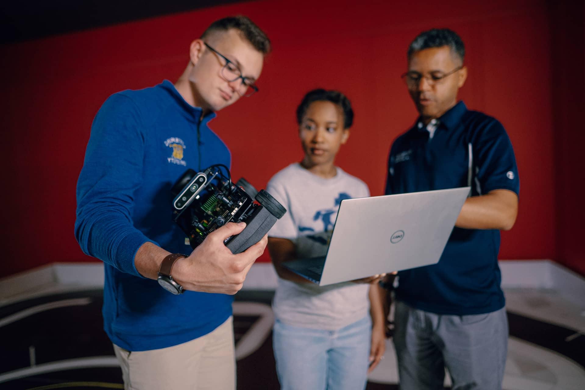 A Kettering professor points at a laptop that a student holds. Another student holds and scrutinizes a self-driving robot.